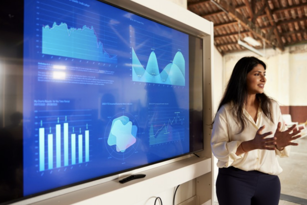 A business professional gives a presentation in front of a screen showing charts and graphics.